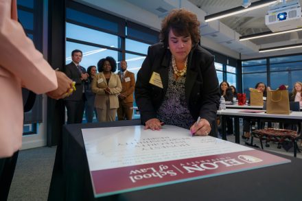 A woman in professional attire uses a marker to sign a poster with the words Honesty, Integrity, Responsibility, Respect. Other students are shown behind her waiting to sign.