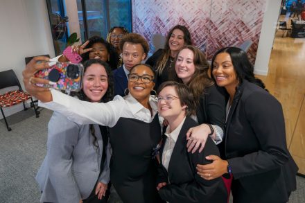 A group of nine students gather for a group selfie photo. One in front has their arm outstretched holding a phone.