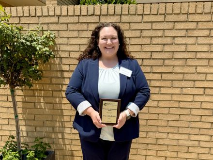 A woman holding an award plaque outdoors. She is wearing a blue suit and smiling at the camera. A brick wall is behind her.
