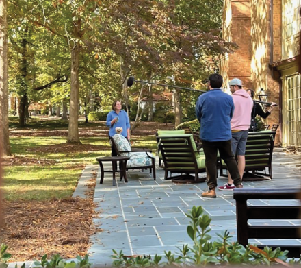 A group of university alumni hold a camera and a boom mic while interviewing a university president outside beneath a large tree.