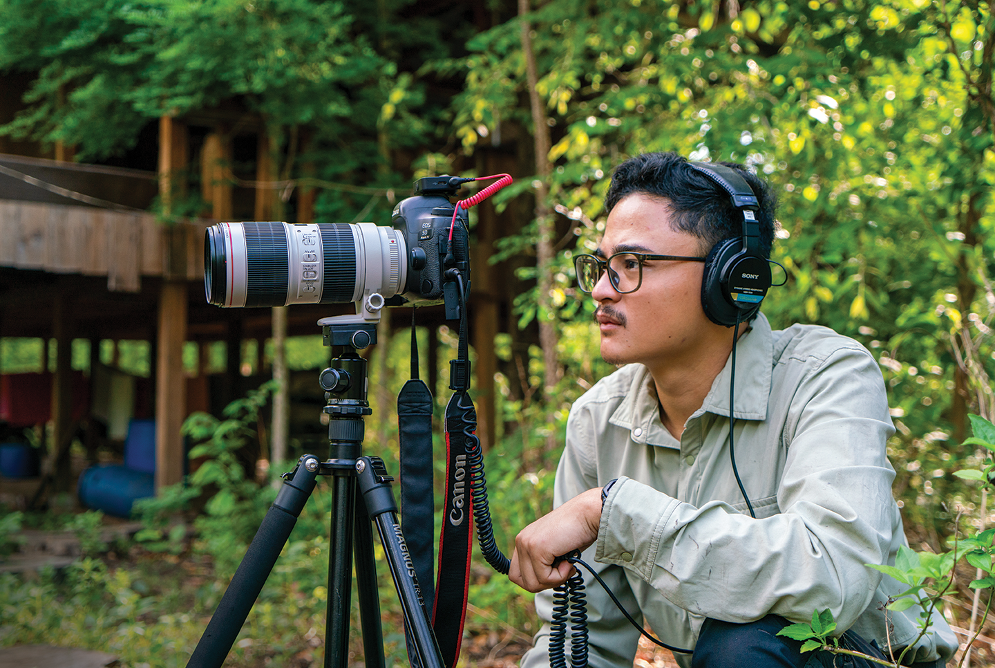 A man wearing headphones crouches next to a camera on a tripod in a forest.