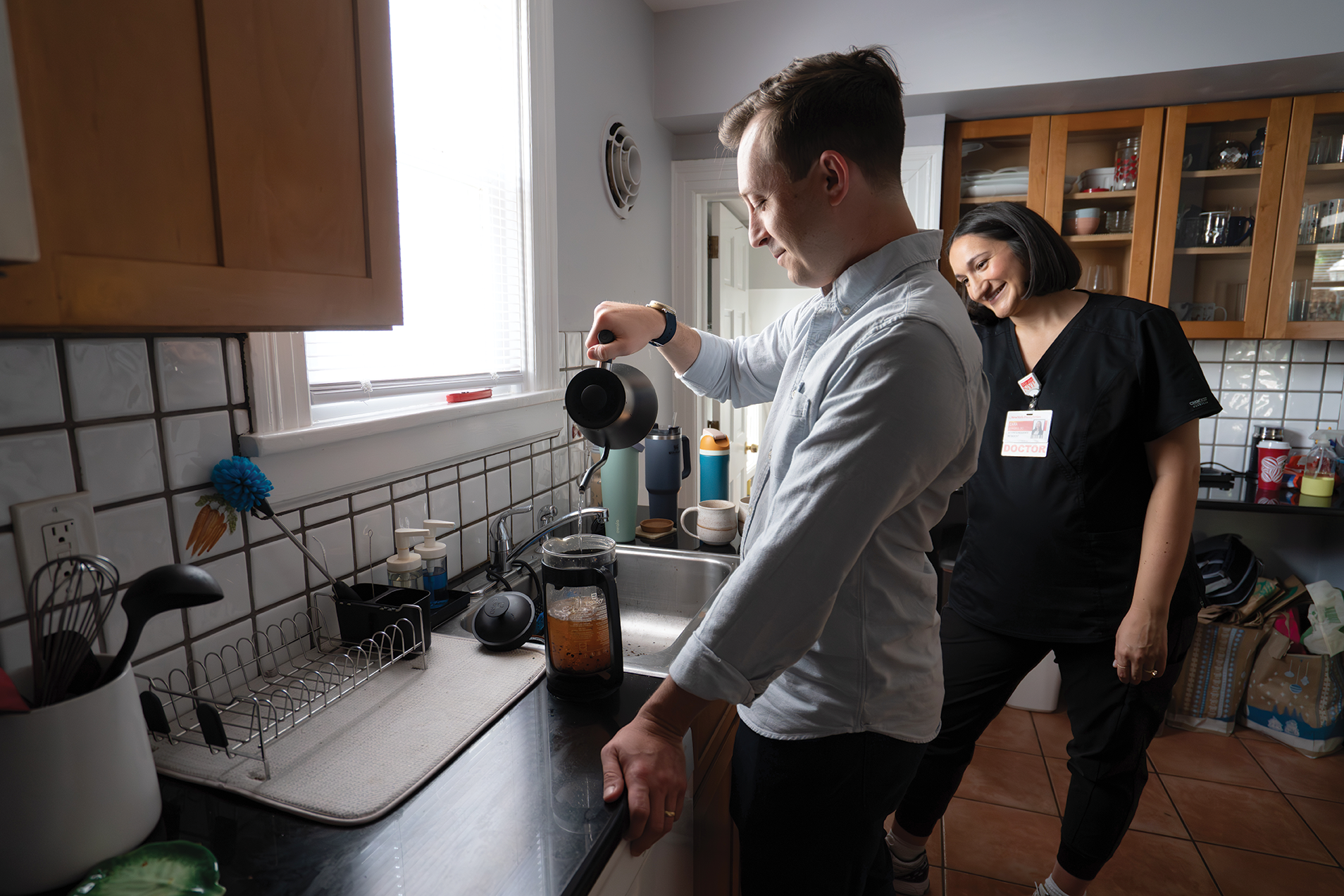 A man stands at a kitchen counter prepares coffee while a woman wearing medical scrubs stands beside him and smiles.
