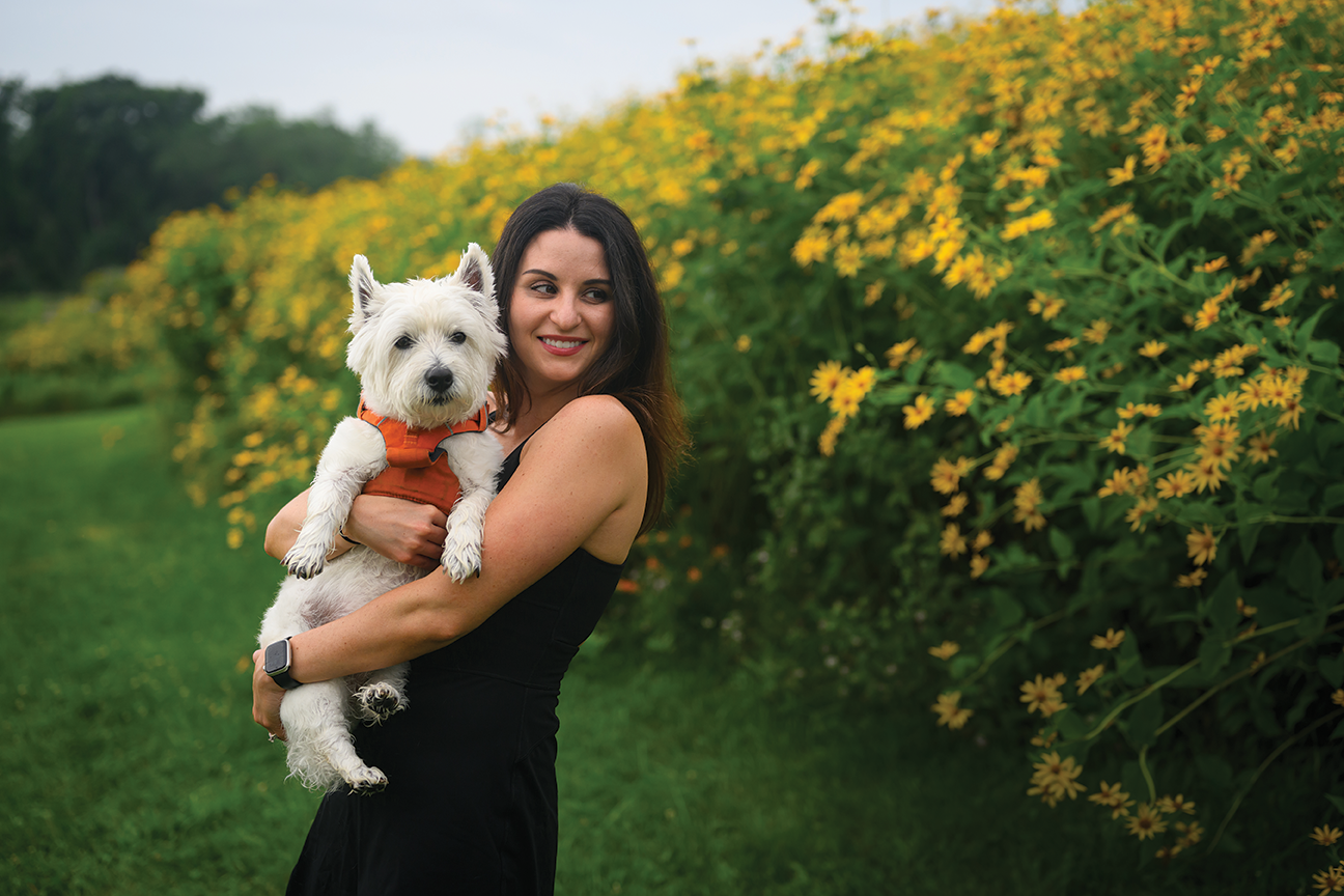 A woman holds a small white dog in a field of flowers.