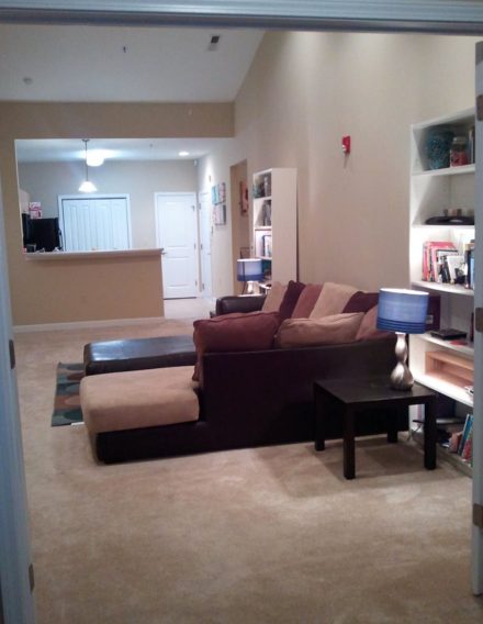 Spacious living room with a sectional sofa and bookshelves, facing an open kitchen and entryway under soft ceiling lights.