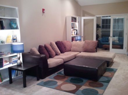 Cozy living area with a large sectional, patterned rug, ottoman, and built-in bookshelves, with French doors in the background.