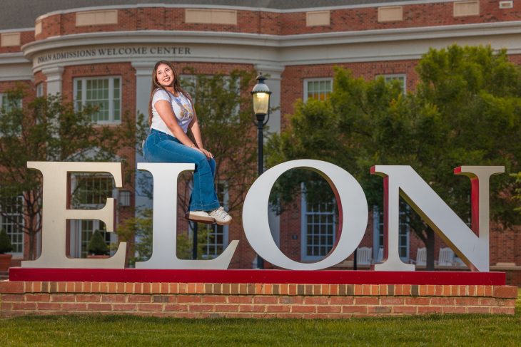 A smiling student poses sitting atop the large ELON letters in front of the Inman Admissions Welcome Center at sunset.