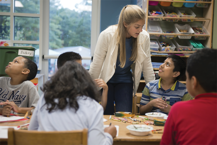 A young boy sits at a table of art supplies with other children. He looks up at a female teacher and talks to her.