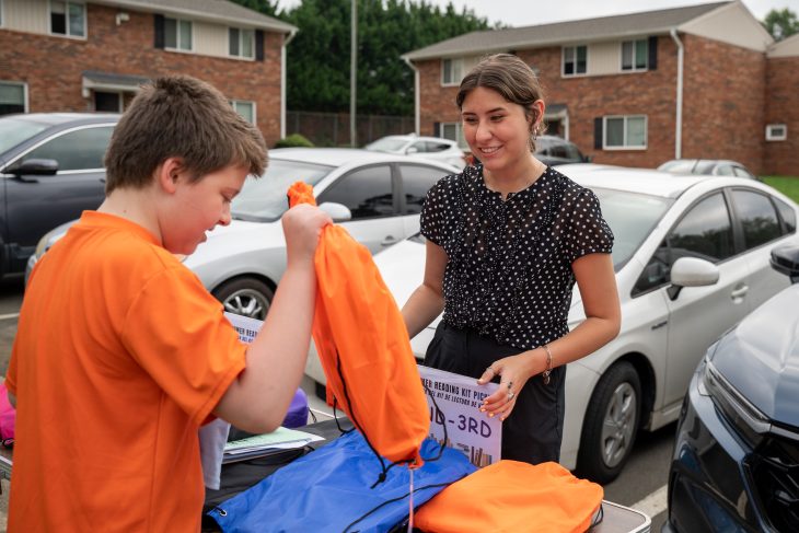 A smiling woman hands an orange drawstring bag to a boy in an orange shirt during an outdoor event in a residential parking lot.