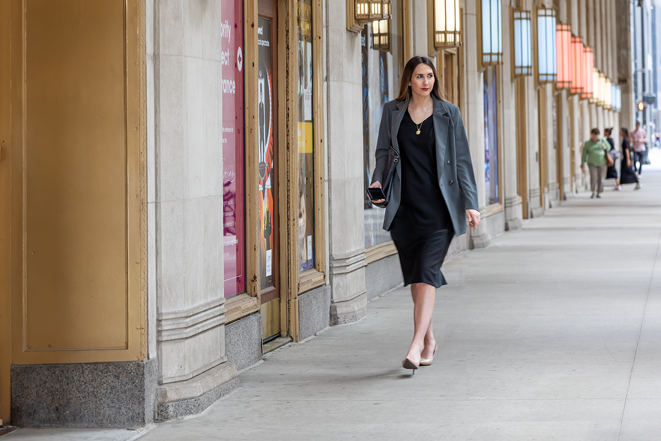 A woman wearing business attire walks down a city sidewalk with her phone in her hand.