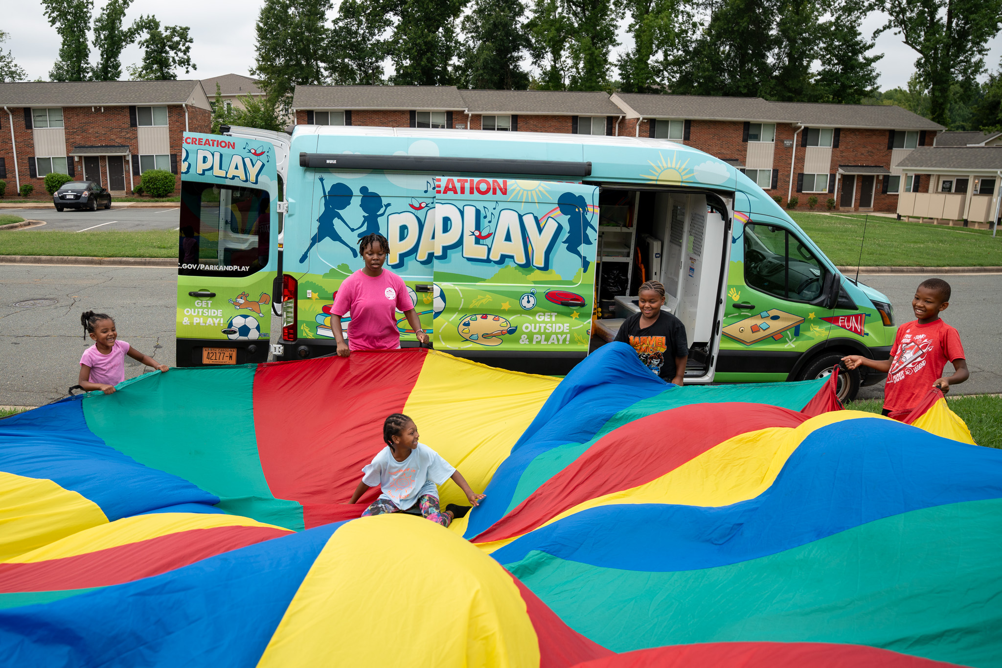 Children play with a colorful parachute in front of a “Pop Play” activity van parked in a residential neighborhood.