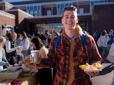 A student smiles while holding a cup of coffee and a plate of fruit and pastries at an outdoor event. Other students gather at tables in the background under bright sunlight.