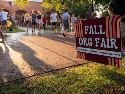 A maroon sign with retro stripes reads “Fall Org Fair, Thursday 8.29.24, 6–8 p.m., Young Commons” as students walk along a brick path in the background during golden hour.