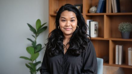 A woman with dark long hair, a dark top and gold earrings sits in front of a bookshelf with various items with a plant next to it.