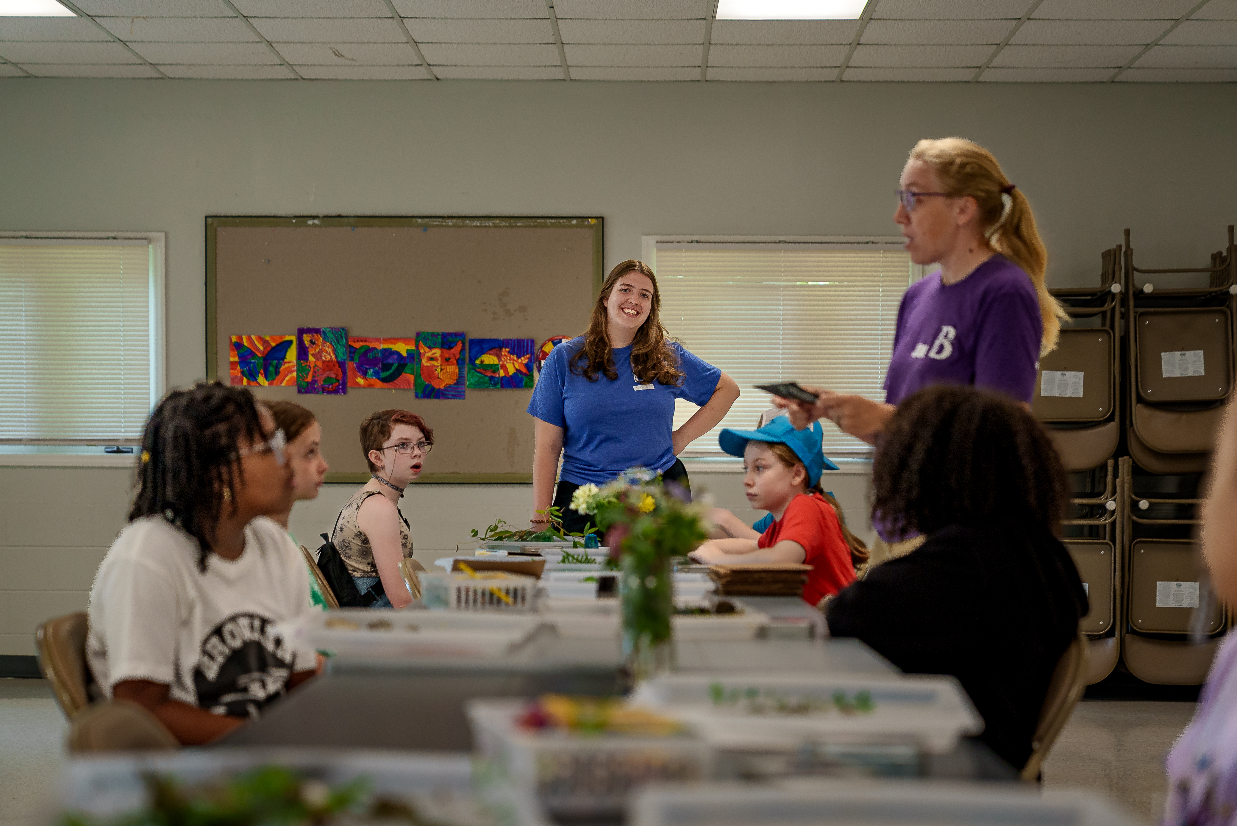 A smiling instructor in a blue shirt stands as campers seated around tables work on a project in a classroom decorated with colorful animal art.