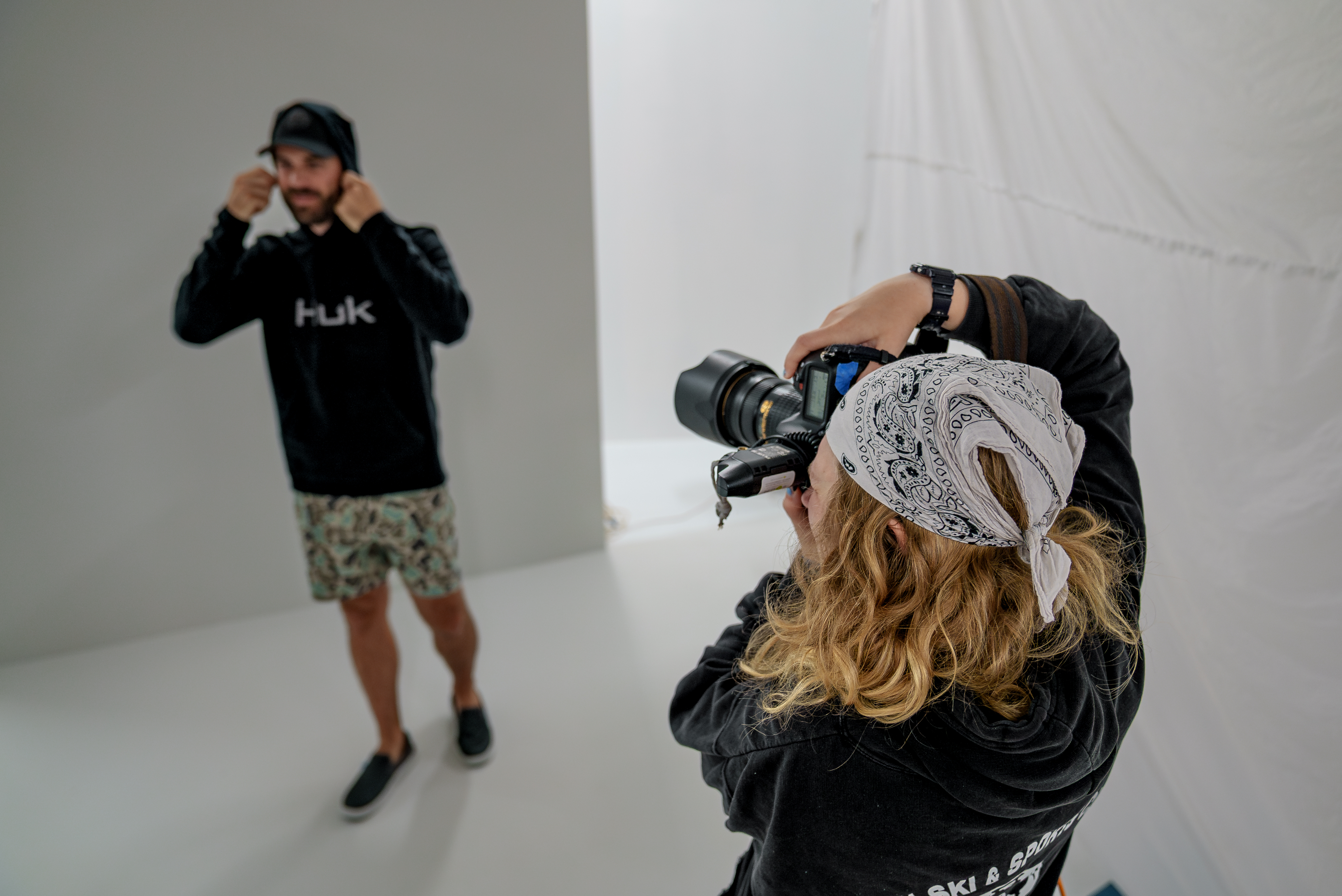 A student photographer wearing a bandana takes a portrait of a model adjusting a hoodie during a photo shoot in a minimalist studio.