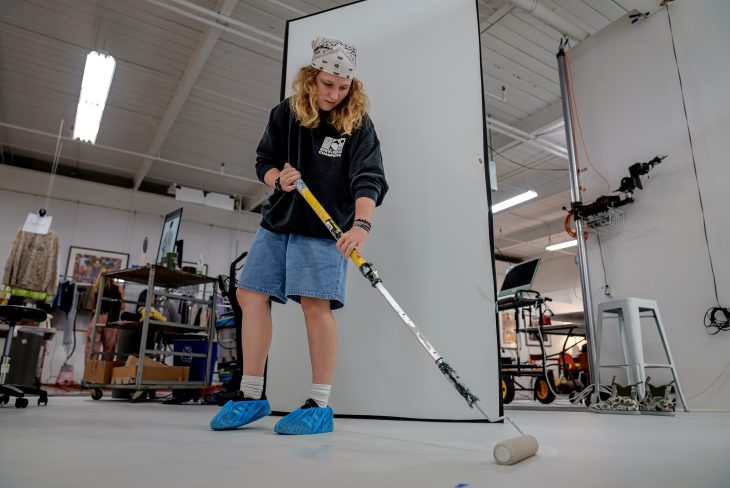 A student paints the floor with a long roller in a photography studio, preparing the space while wearing blue shoe covers and casual clothes.