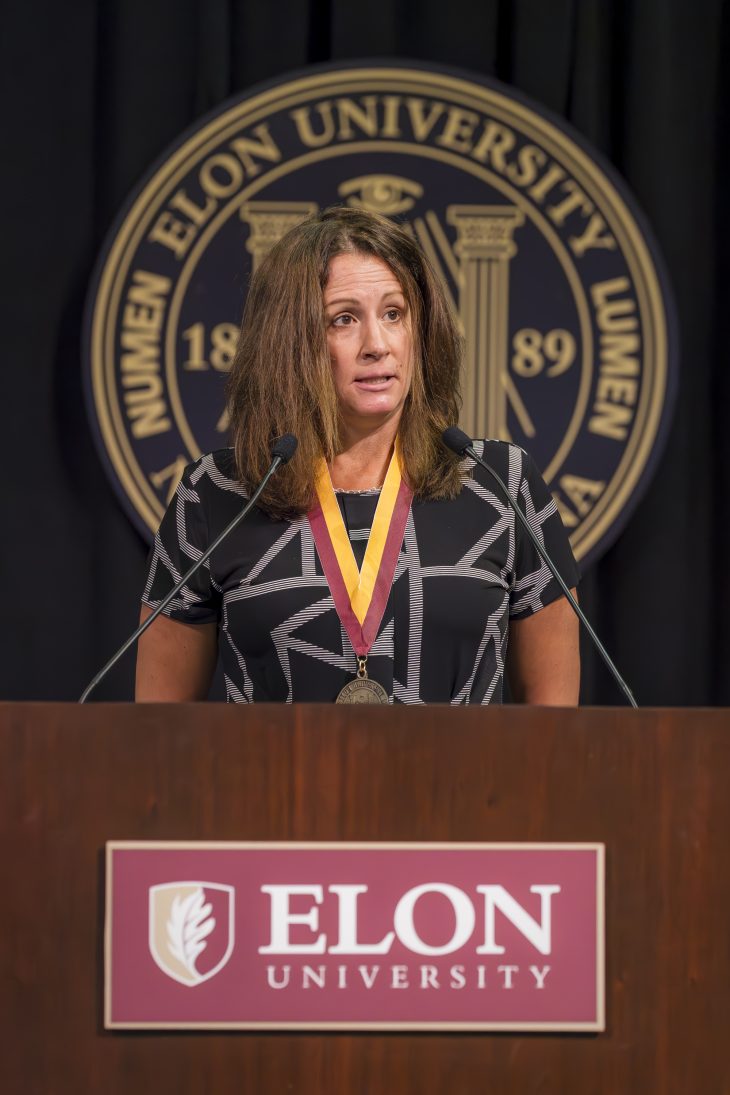 A woman speaks at the podium with the Elon Univesity logo below her and the seal blurred behind her on a black background