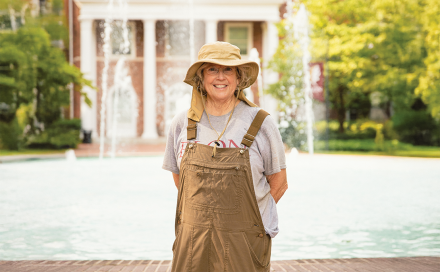 A woman wearing overalls and a gardening hat stands in front of a fountain and smiles