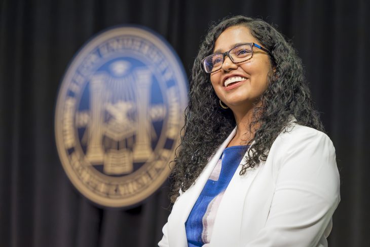 A woman smiles in front of a black backdrop and the Elon seal