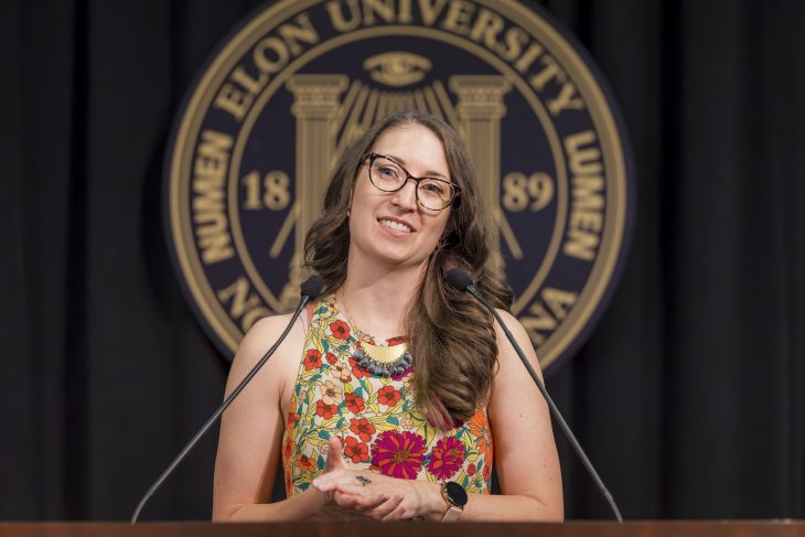 A woman speaks at the podium with the Elon Univesity logo below her and the seal blurred behind her on a black background