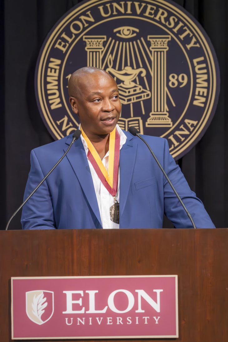 A man speaks at the podium with the Elon Univesity logo below her and the seal blurred behind him on a black background