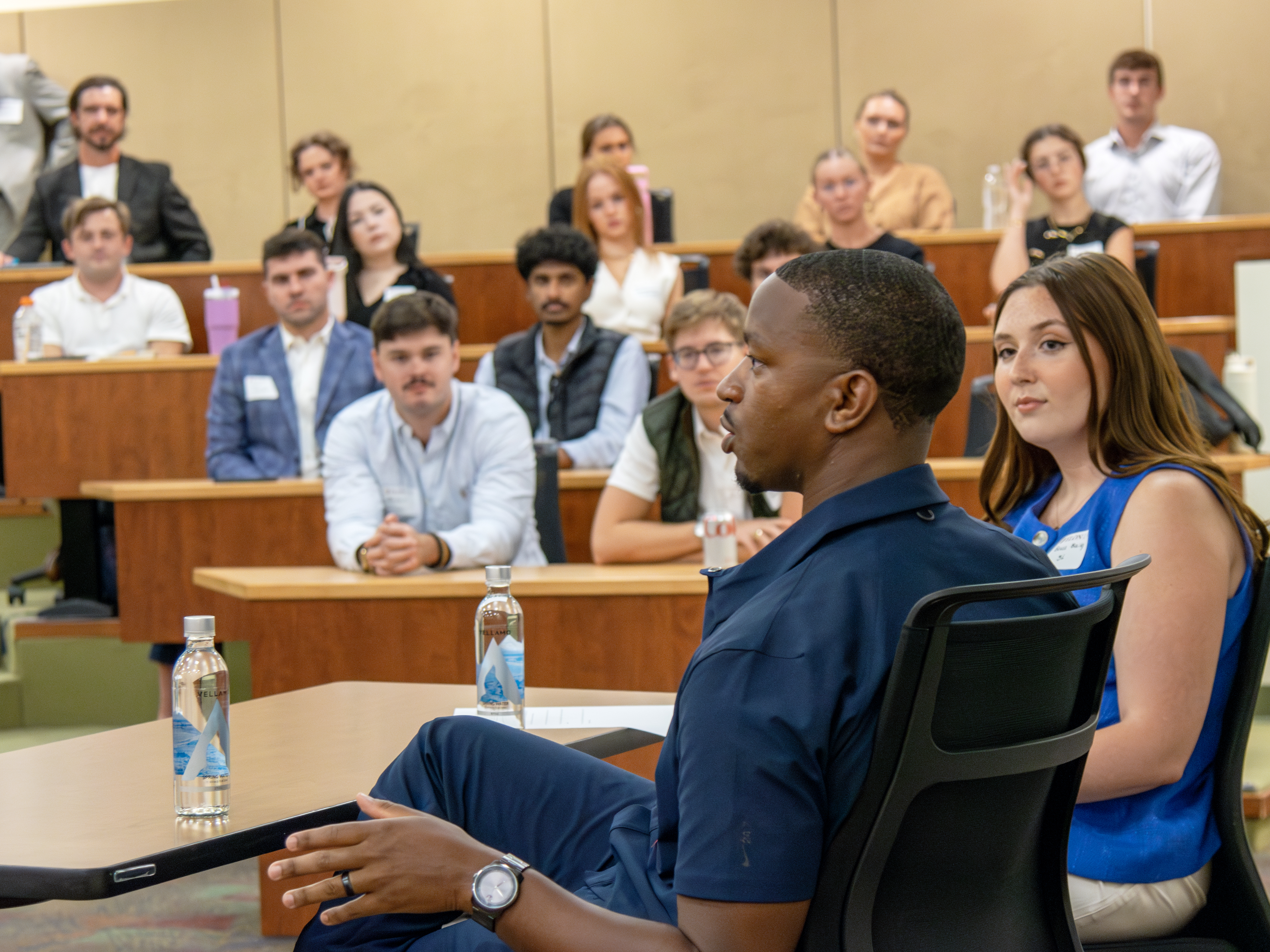 Two people seated in front of a crowded classroom. Students are seen listening attentively in the background.