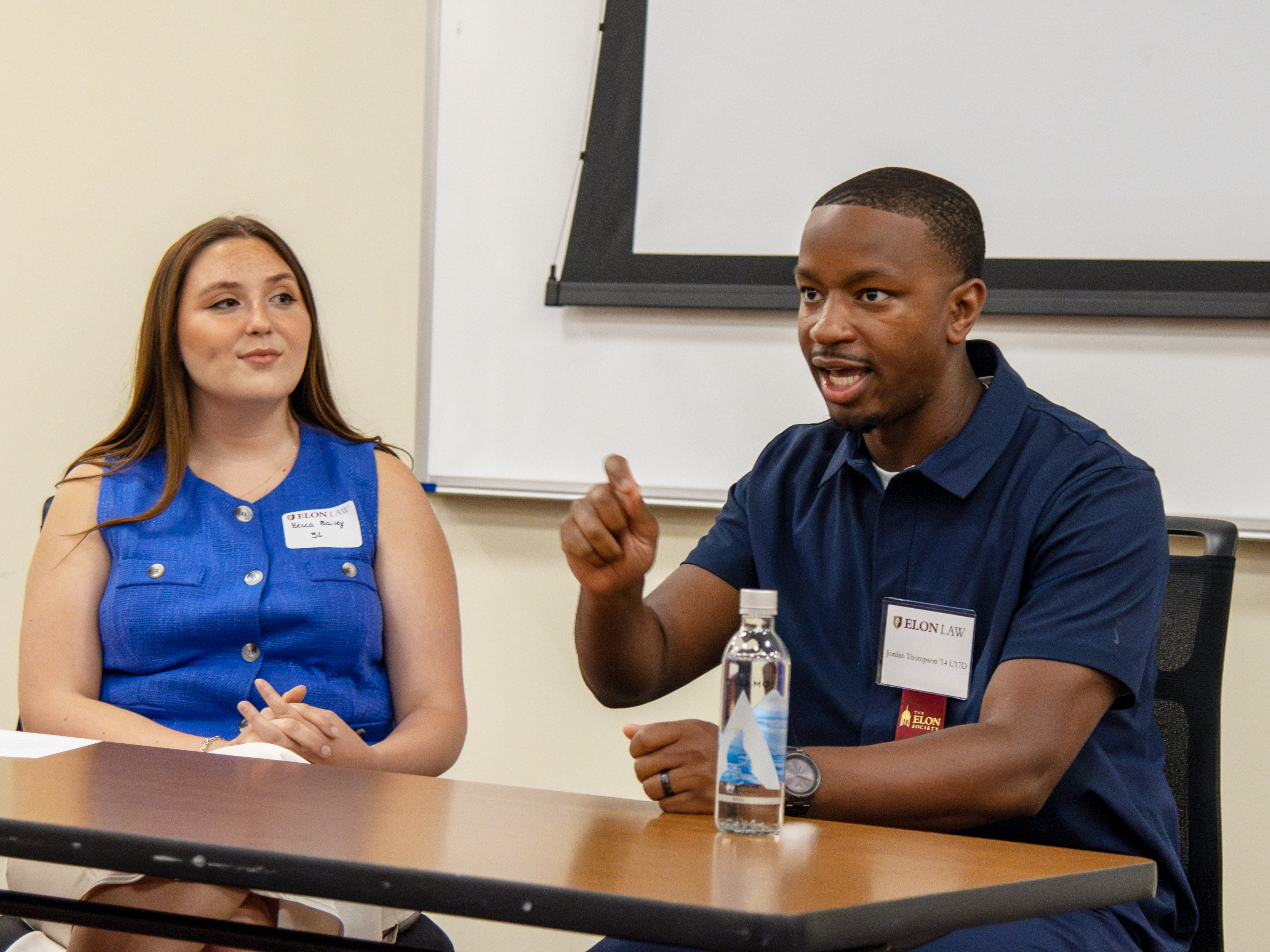 Two people seated at a table. A man is gesturing with his right hand. The woman looks on smiling.
