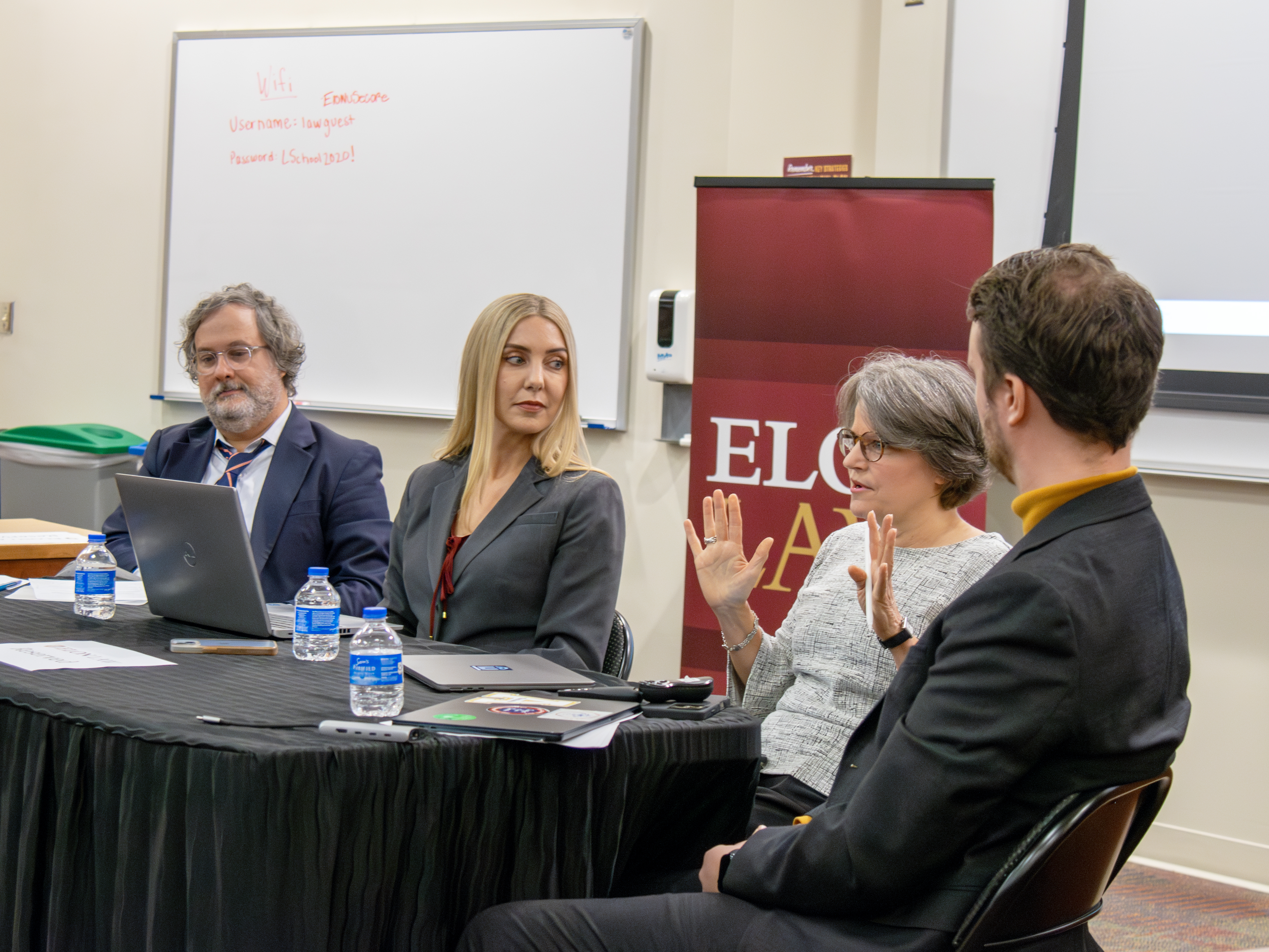 Four people seated a table in a large classroom. A banner with "Elon Law" is shown behind them.