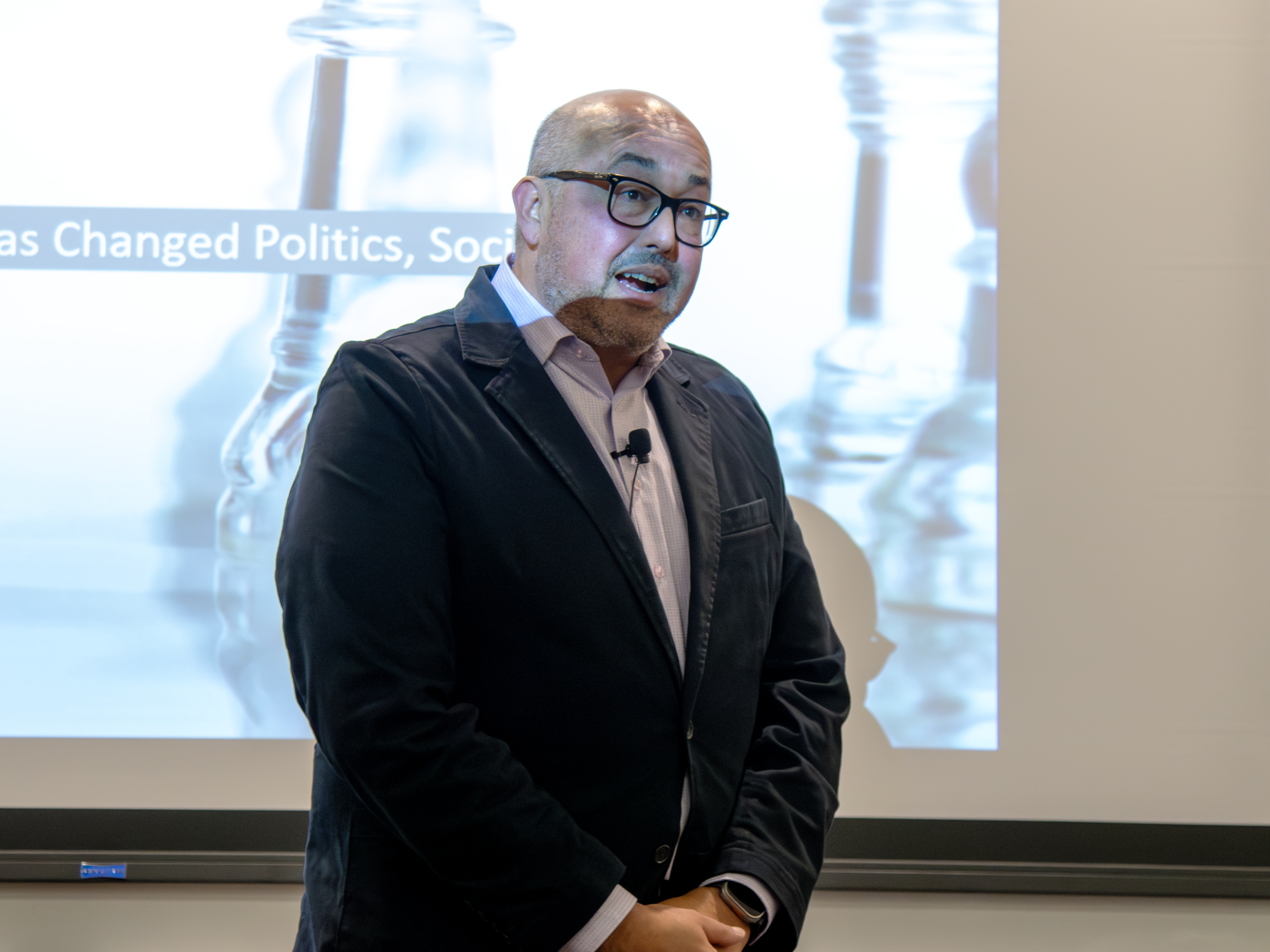 A man stands speaking in front of a projected image of chess pieces.