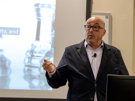 A man gestures while speaking at a podium. Images of chess pieces are projected on a screen behind him.