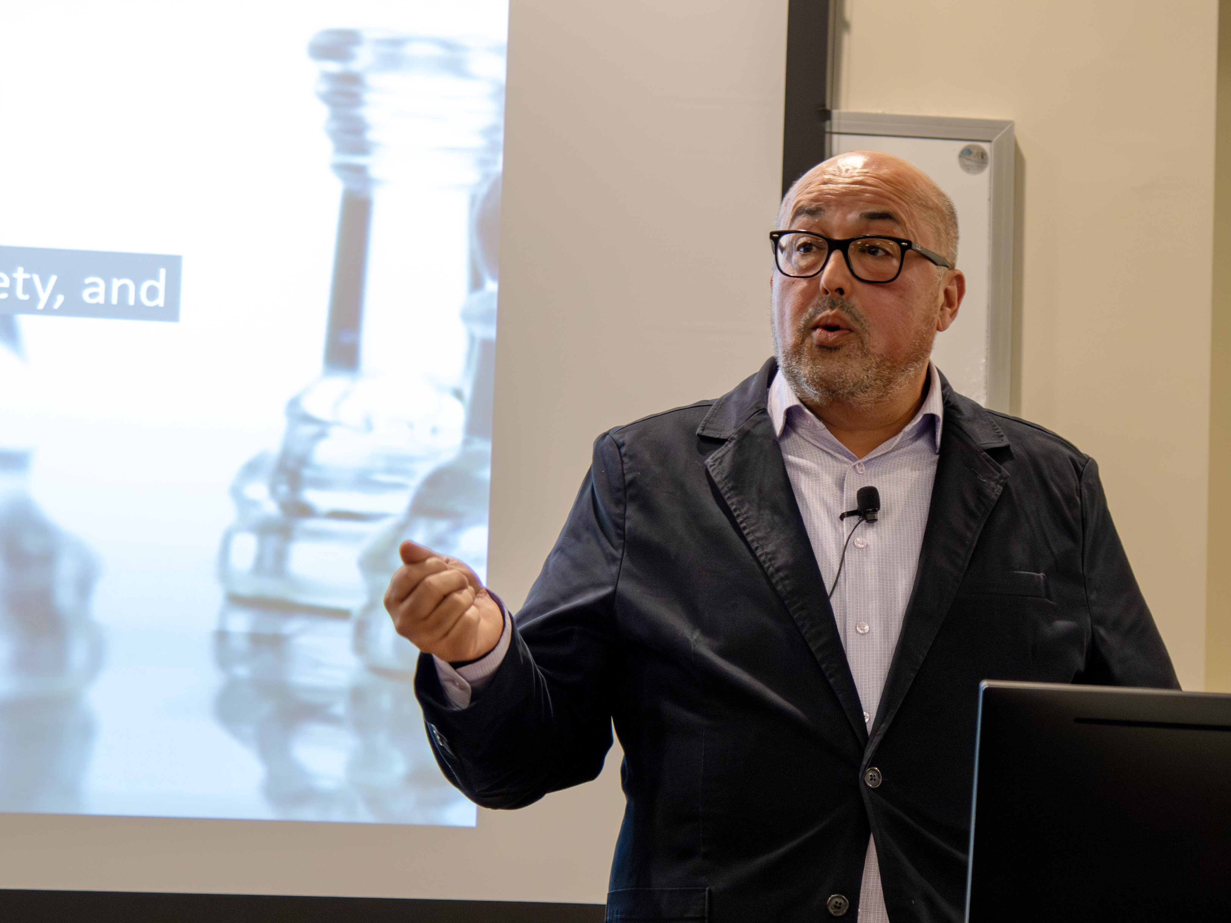 A man gestures while speaking at a podium. Images of chess pieces are projected on a screen behind him.