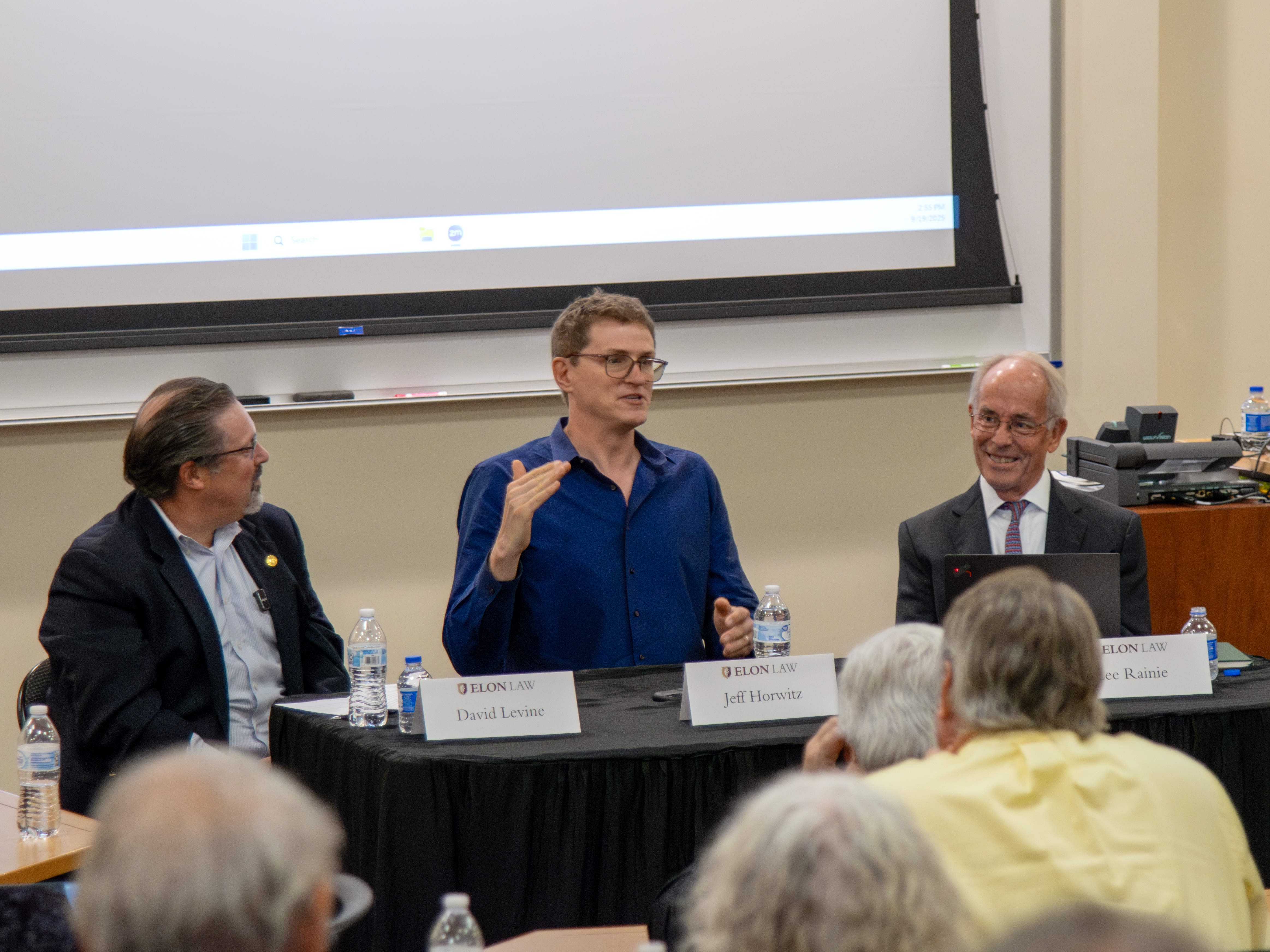 3 men seated at a table in a classroom. Two are listening to the man in the center speak.