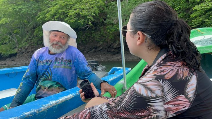 A student holds a voice recorder while interviewing someone on a boat.