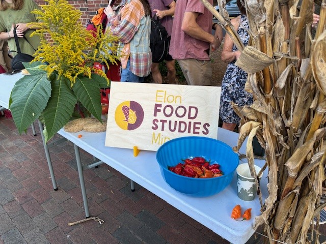 Table with Elon Food Studies sign, blue bowl of peppers, yellow flowers, and cornstalks at an outdoor event.