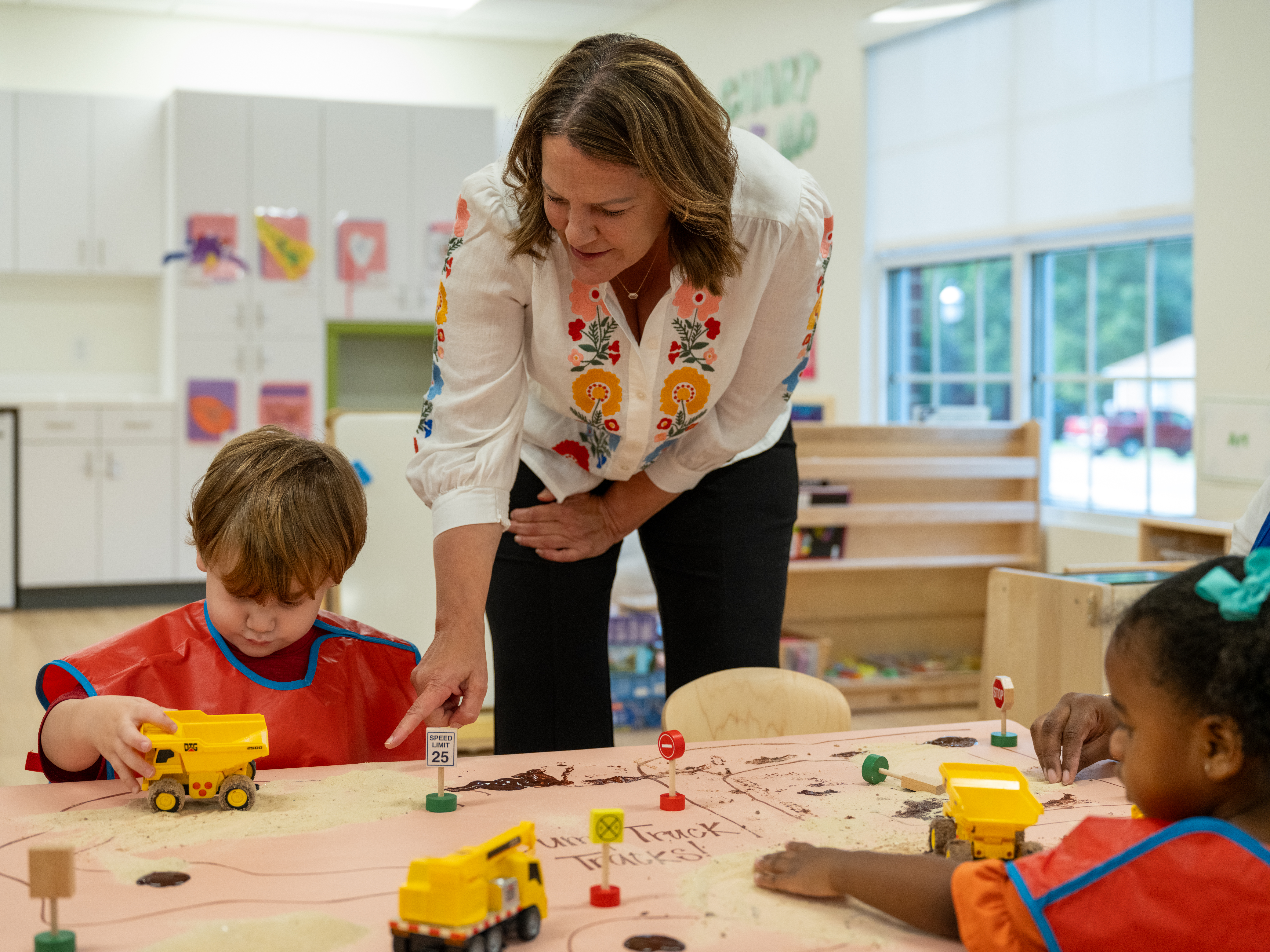 A woman with medium brown hair points at a toy dump truck that a child in a red apron is playing with.