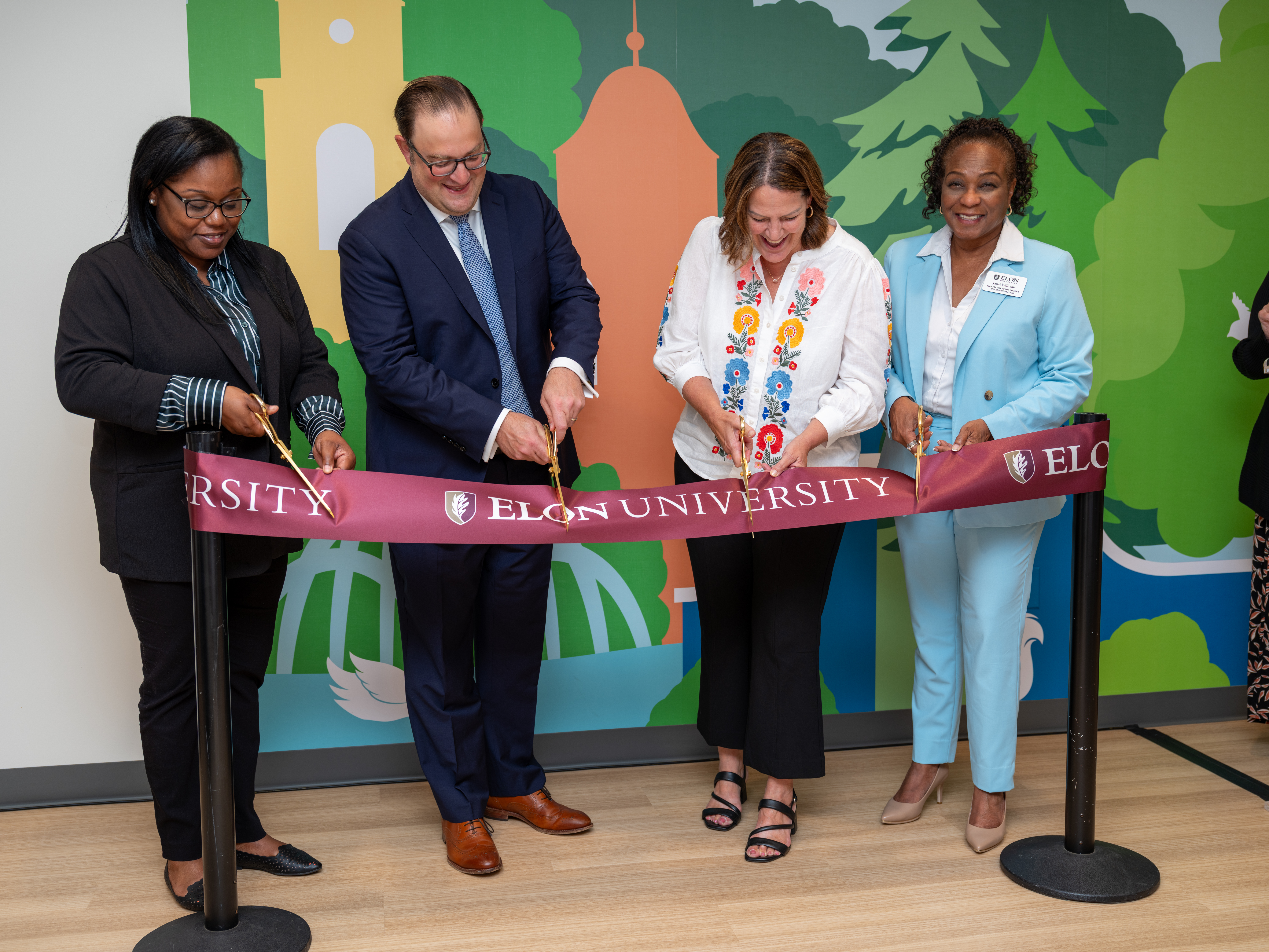 Four people stand on a light wooden floor with golden scissors in their hands. They cut a maroon ribbon with Elon branding. A colorful mural is in the background featuring a forest scene.
