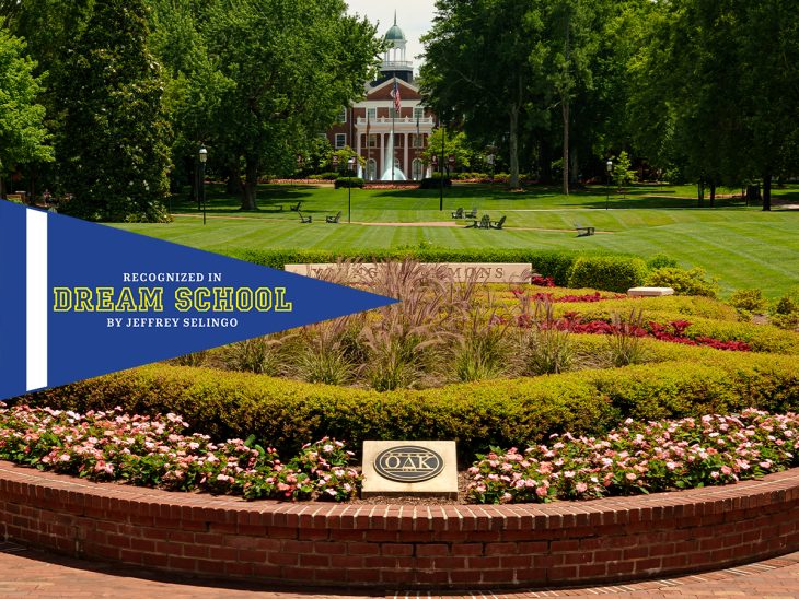 An view of a beautifully landscaped university campus with a central brick pathway, manicured lawns, colorful flowerbeds, and a stately red-brick building in the background, featuring a banner that reads “Recognized in Dream School by Jeffrey Selingo.”