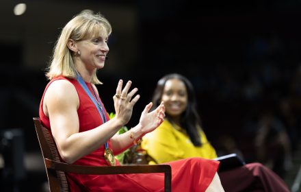 Katie Ledecky, wearing a red dress and gold medal, speaks on stage at Elon University while Zora Stephenson listens in the background.
