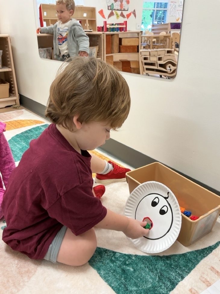 A boy with short hair and red shirt plays with a paper plate with a face drawn on it and a hole for a mouth.