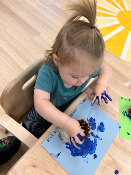 A little girl with a teal shirt sits in a wooden chair and plays with blue paint on a blue piece of paper using a pinecone. 