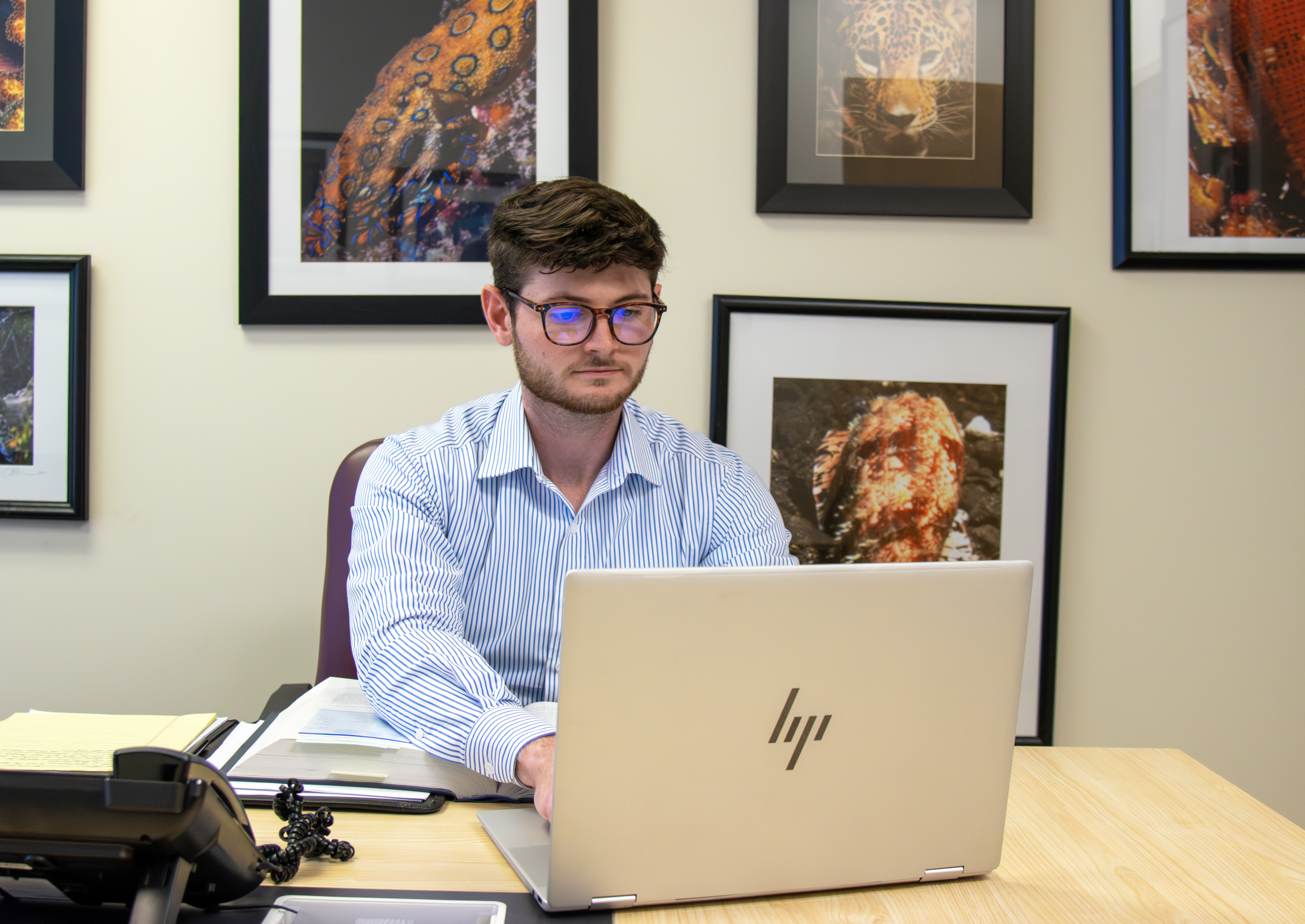 Joshua Burgan sits at a desk typing on a laptop. He is wearing glasses and a blue shirt. Nature photos hang on the wall behind him.