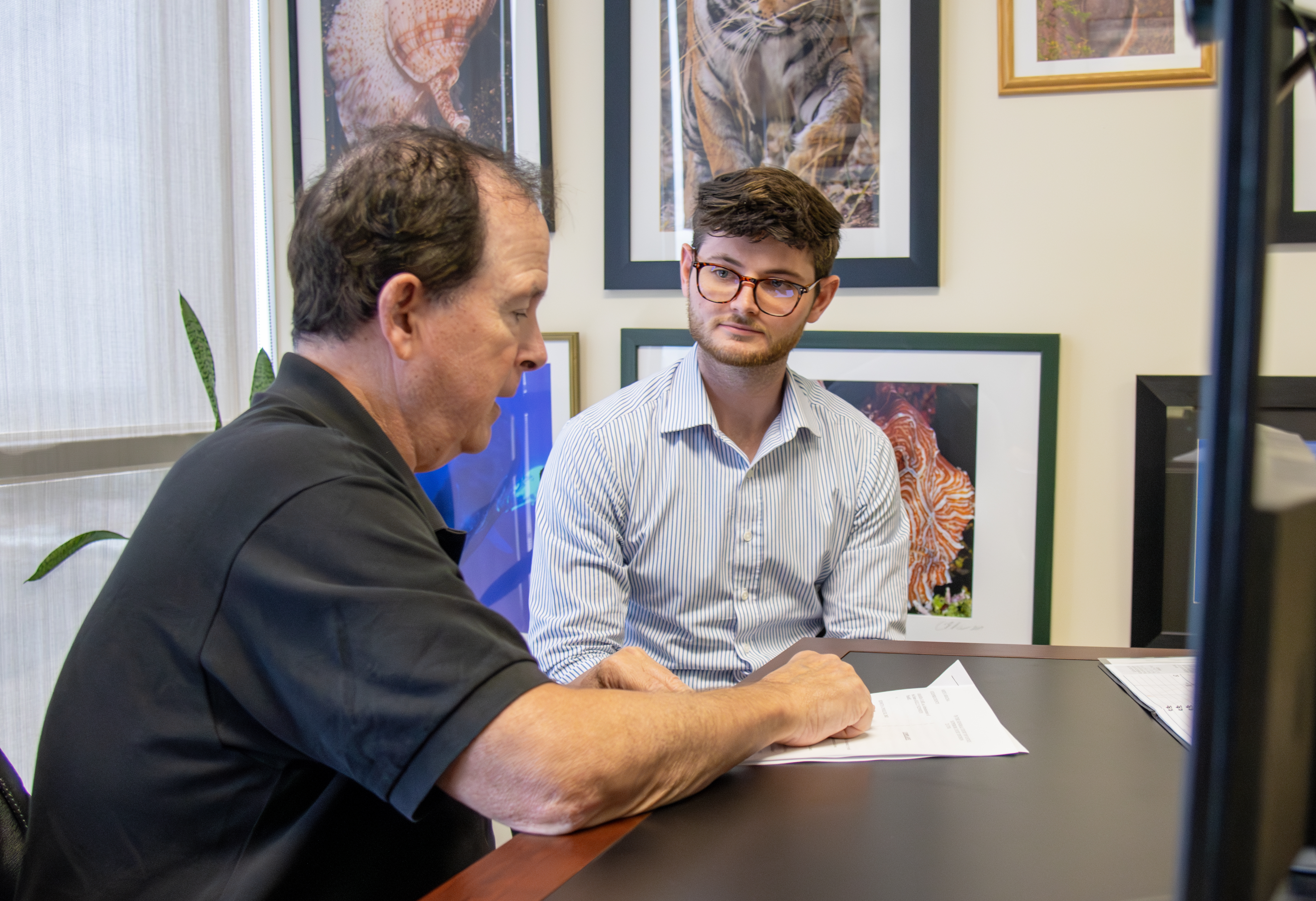 Two men seated at a desk. One man speaks and points to a document. The other is listening intently.