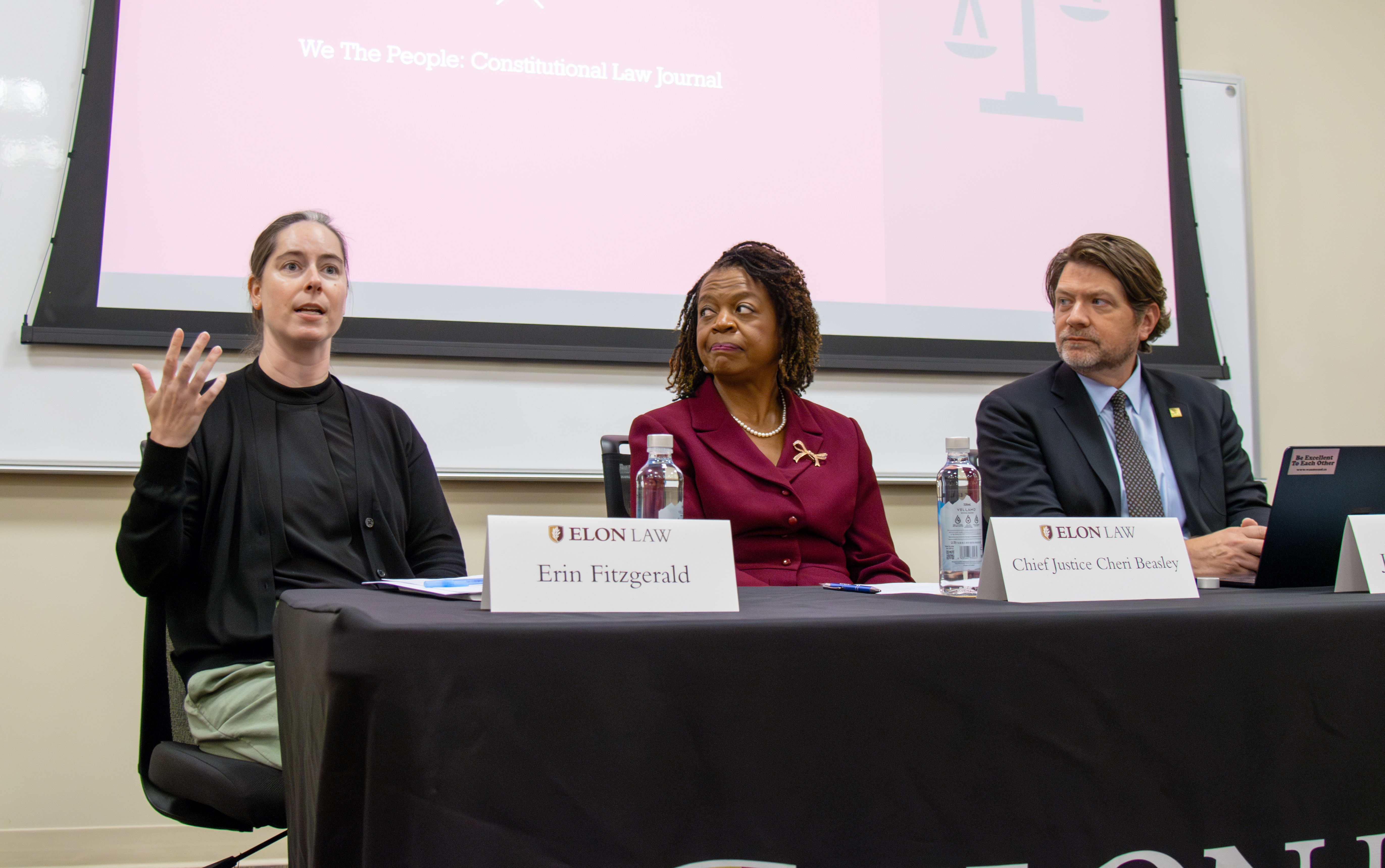 Three people seated behind a table with name plaquards in front of them. The woman on the left is gesturing while speaking.