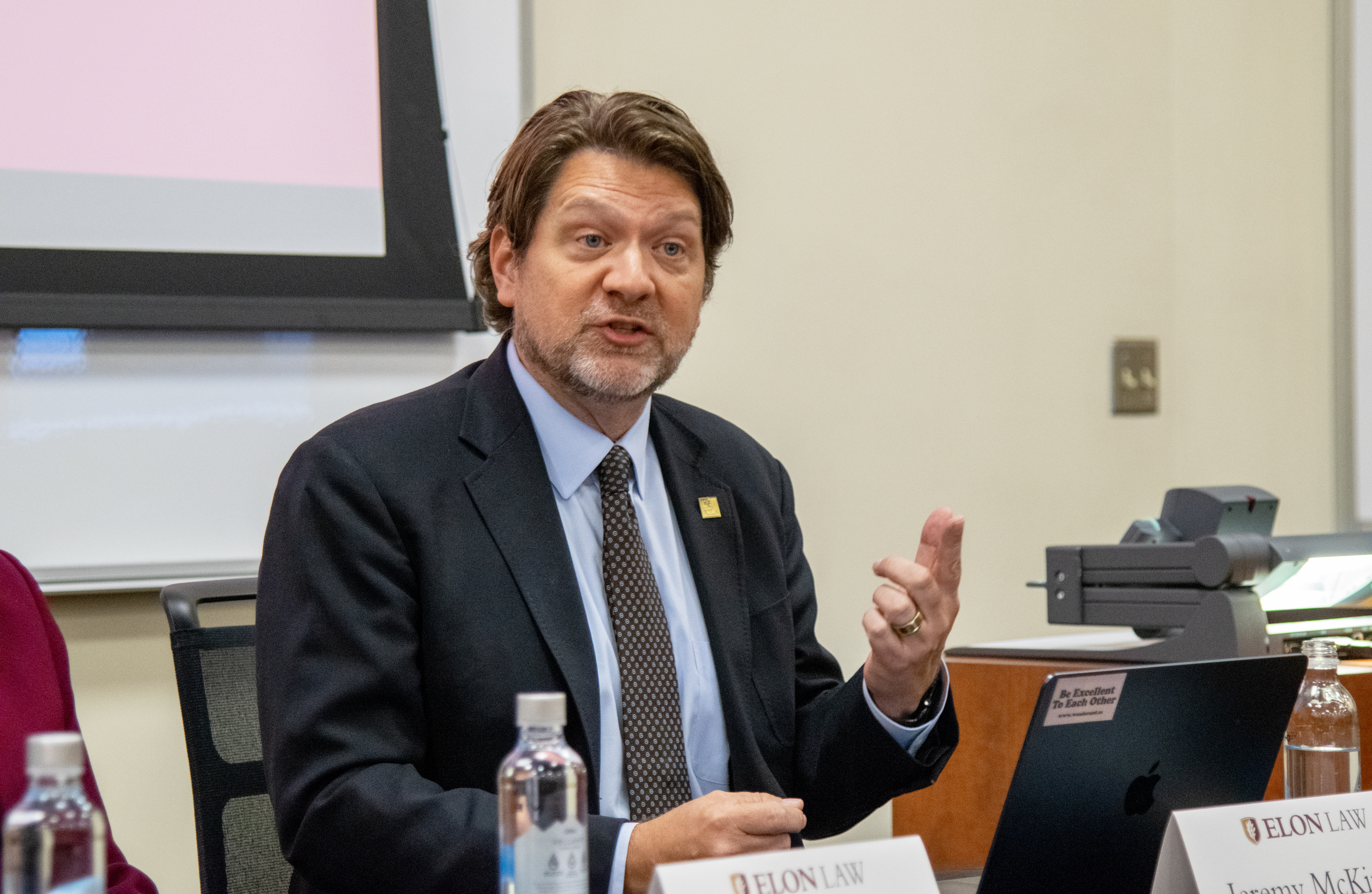 A man gestures while speaking seated behind a table.