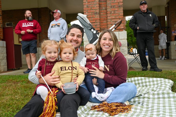 A family sits on a picnic blanket wearing Elon University gear