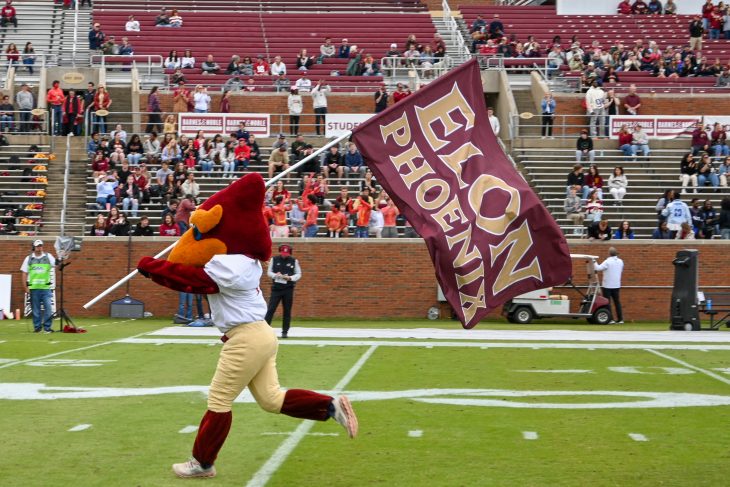 The Elon University Phoenix mascot runs across the football field carrying a large Elon Phoenix flag.