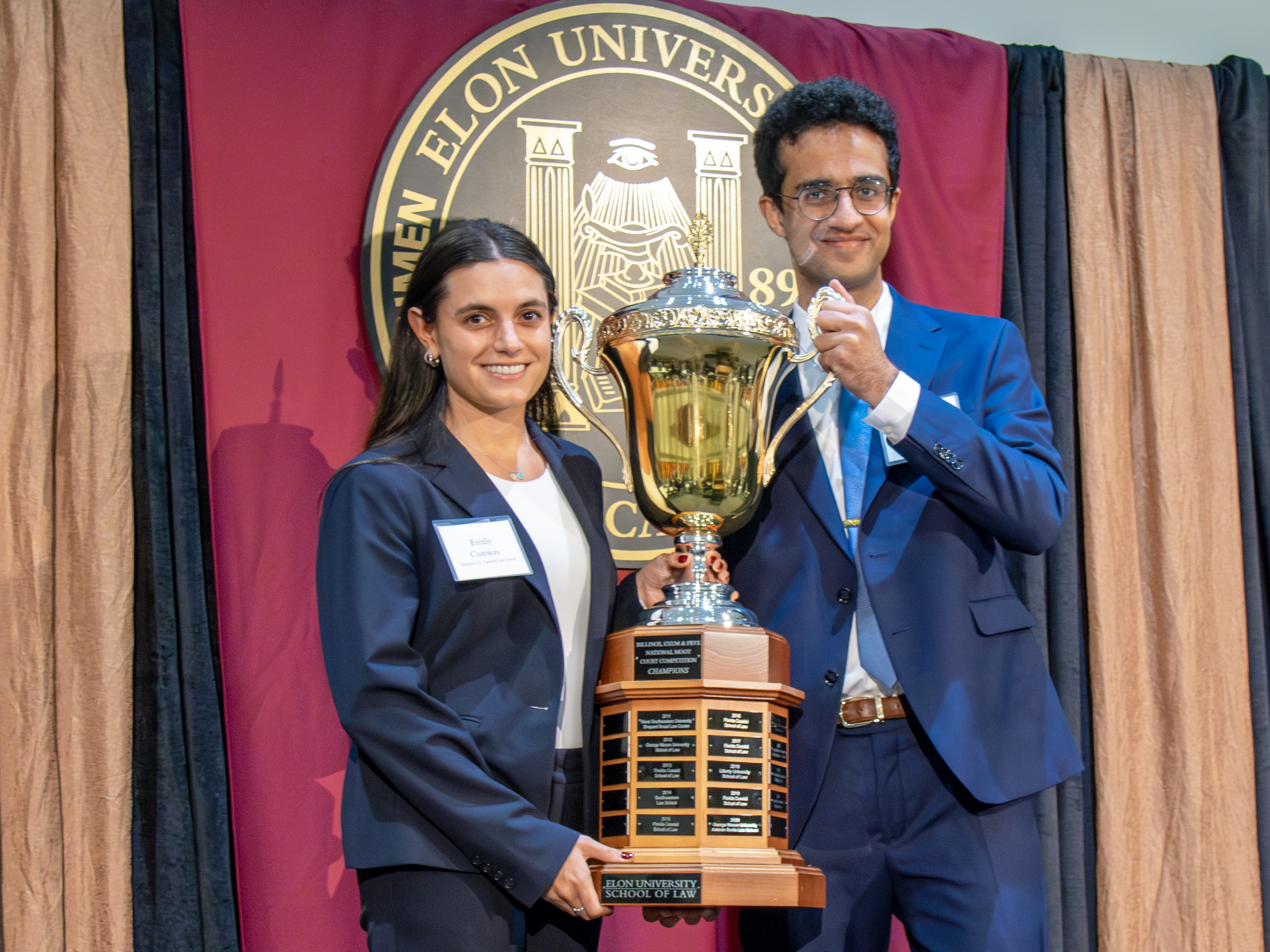 A man and woman hold a large trophy cup on a stage with the Elon University seal behind them