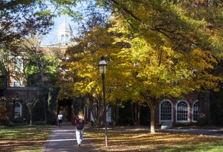 A student walks along a brick path lined with trees showing bright yellow fall leaves, with a historic red-brick campus building and clock tower in the background.