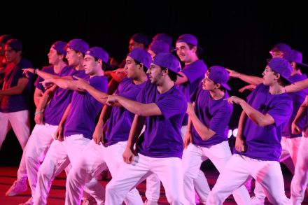 A group of students performs a synchronized hip-hop dance on stage, wearing matching purple shirts, white pants, and backward purple caps under pink and purple stage lighting.