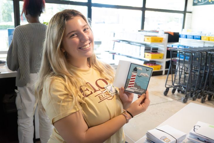 Jenna Toms ’26 smiles while holding a handmade card featuring a red-and-white striped lighthouse and the words “shines brightest” during a volunteer project at Classroom Central. Stacks of cards and school supplies are visible on the table beside her.